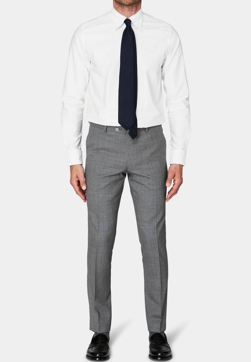 Camicia bianca con colletto, abbinata a pantaloni grigi a scacchi e una cravatta blu scuro; le scarpe sono in pelle nera. Taglio su misura.