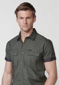 Camisa de manga corta con botones en verde oliva con ribetes de mezclilla oscura, bolsillos en el pecho y detalles impresos. Tejido ligero con una textura suave.