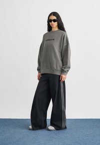 Φαρδιά γκρι φούτερ με μαύρο λογότυπο "adidas", σε συνδυασμό με φαρδιά μαύρα τζιν παντελόνια με γκρι πλευρικές ρίγες, φορεμένα με γυαλιά ηλίου.