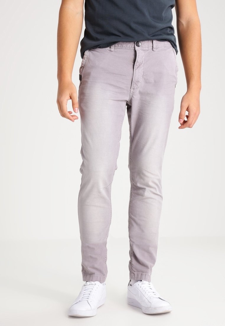 Personne portant un pantalon gris clair ajusté, un t-shirt noir et des baskets blanches, debout devant un arrière-plan uni.