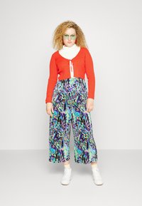 Cardigan curto vermelho com frente amarrada, gola alta branca e calças largas com um padrão abstrato multicolorido. Ténis brancos.
