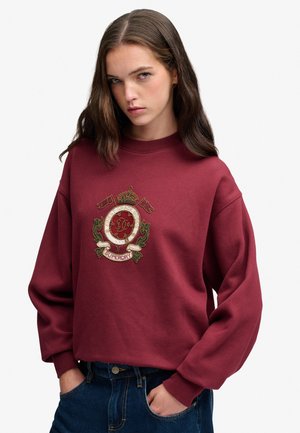 Ung kvinde med langt brunt hår iført en vinrød sweatshirt med et broderet våbenskjold og mørkeblå jeans, hænderne i lommerne.