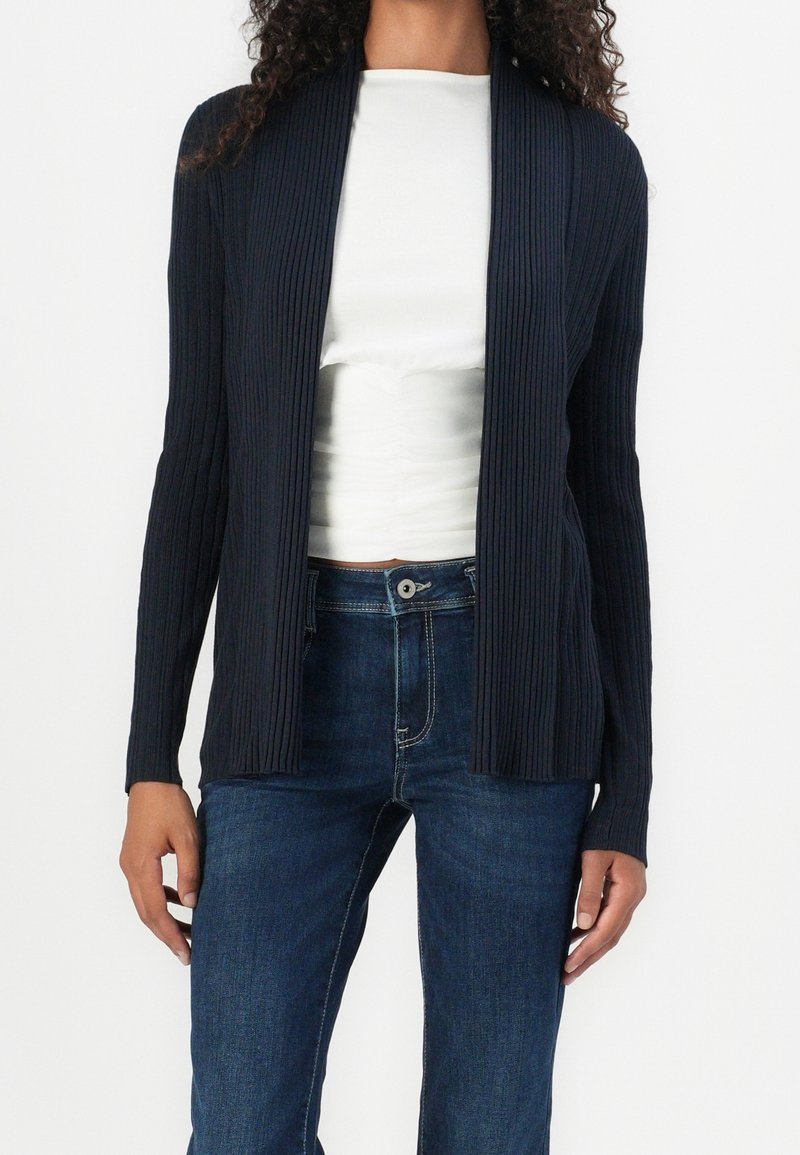 Femme portant un cardigan noir côtelé ouvert sur le devant, par-dessus un top blanc, associé à un jean bleu foncé taille haute.