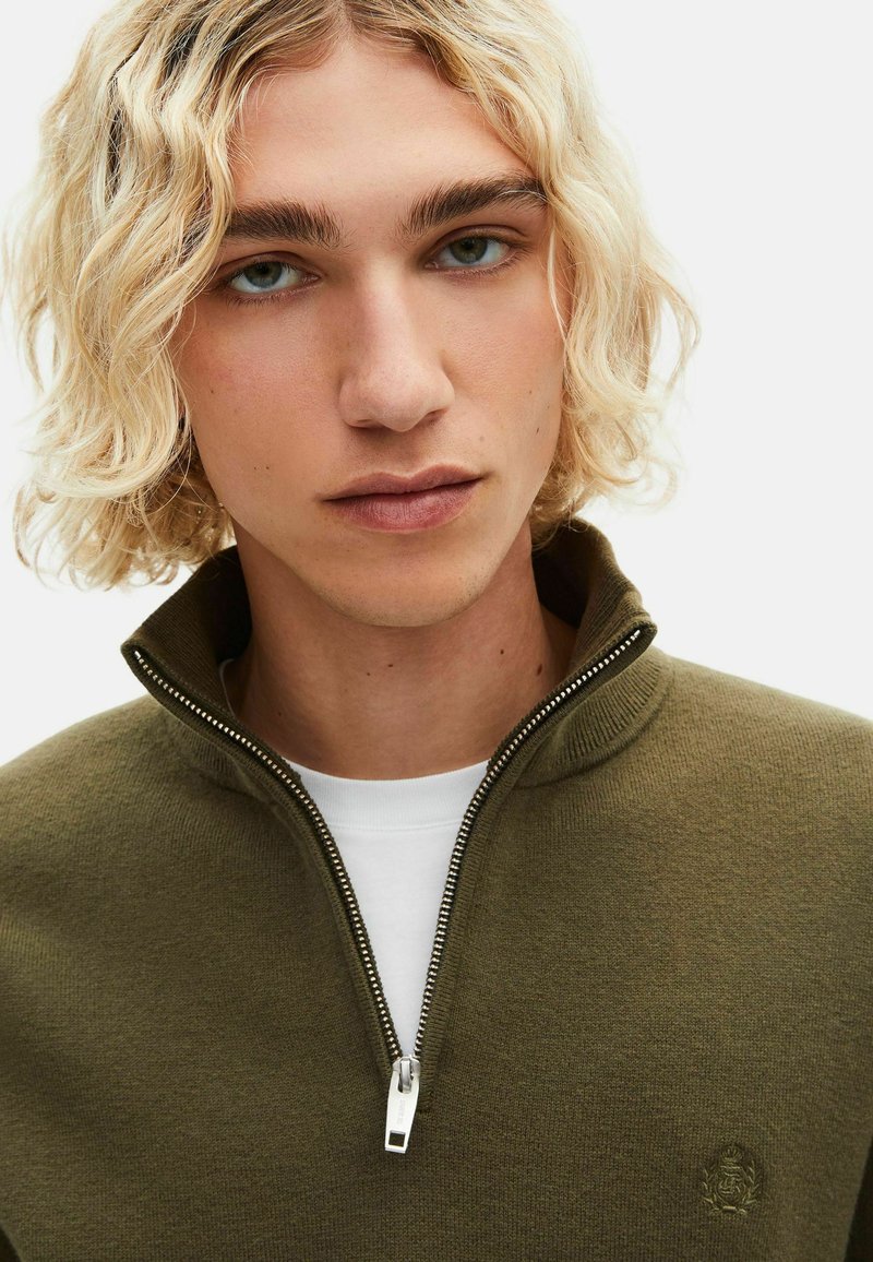 Maglione pullover verde oliva con zip, realizzato in tessuto morbido, con colletto alto e un piccolo emblema sul lato sinistro del petto.