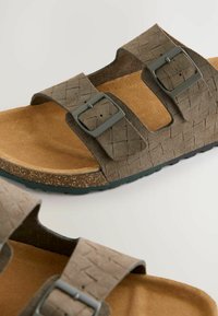 Sandales en suede marron avec deux bandes texturées, boucles métalliques ajustables et une semelle en liège. Caractéristiques d'une semelle extérieure en caoutchouc pour une bonne traction.