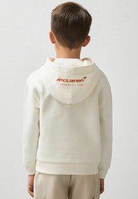 Kremna kapuca z kengurujskim žepom, z ribanim manžetama in robom; na hrbtu je oranžni logotip McLaren. Narejena iz mehkih materialov.