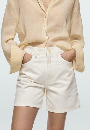 Frau trägt ein hellbeiges Leinenhemd mit Knöpfen und langen weiten Ärmeln, das in weiße, hoch taillierte Denim-Shorts gesteckt ist, Hände in den Taschen, silberne Anhängerkette.