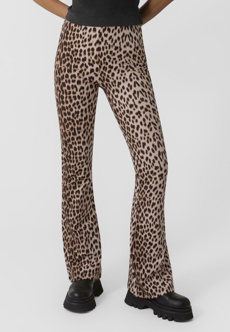 Leopardmönstrade utsvängda byxor i beige och brun, tillverkade av mjukt tyg. Har en åtsittande midja och design med utsvängda ben, kombinerat med svarta, kraftiga stövlar.