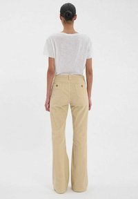 Pantalons en velours côtelé beige évasés avec une taille haute, dotés de deux poches arrière et de boutons en laiton, associés à un T-shirt blanc simple.