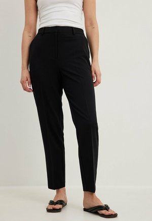 Vrouw draagt strakke zwarte broek, wit mouwloos topje en zwarte sandaal met knoopdetail, staand tegen een effen lichte achtergrond.