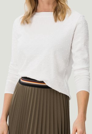 Femme portant un chemisier blanc texturé à manches longues, rentré dans une jupe plissée taille haute vert olive avec une ceinture rayée.