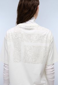 Witte T-shirt met een textuurgrafiekontwerp op de achterkant in licht beige. Oversized pasvorm met korte mouwen en een ronde hals.