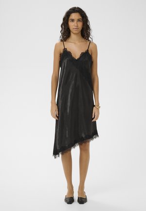 Femeie purtând o rochie neagră asimetrică din satin, cu dantelă la margine și pantofi negri cu vârf ascuțit, stând în picioare pe un fundal alb simplu.