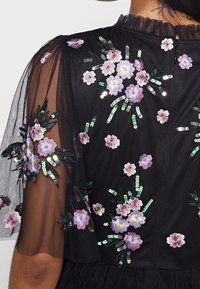Robe noire ornée de broderies florales en violet et blanc, avec des sequins verts. Manches transparentes avec des bords volants, tissu texturé.