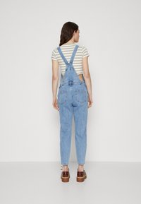 Pantalones de mezclilla en un tono azul claro, con tirantes ajustables, pierna ceñida y dos bolsillos traseros. Llevados sobre una camiseta corta de rayas con mangas cortas.