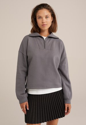 Svetlošedá mikina na zips s vysokým golierom a voľným strihom; má ribované manžety a čistý, minimalistický dizajn. Pod ňou je čierna plisovaná sukňa.