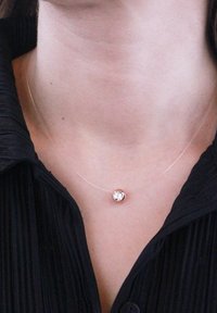 Collana sottile e trasparente con un pendente rotondo in oro rosa, che presenta al centro una pietra trasparente e scintillante. Indossata sopra una maglia scura e texturizzata.