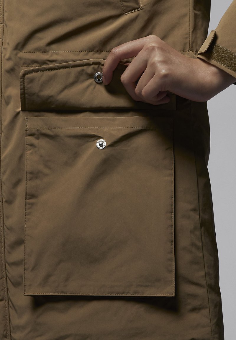 Chaqueta impermeable marrón con un gran bolsillo frontal que cuenta con un cierre a presión. La tela tiene una textura suave y un diseño sencillo.