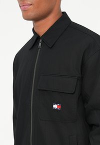 Mężczyzna w czarnej kurtce zapinanej na zamek z kieszenią na klatce piersiowej i logo Tommy Jeans na tle białym.