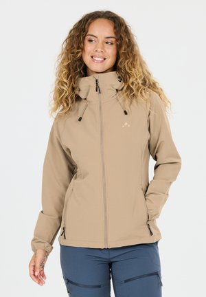 Frau mit lockigem Haar, die eine beigefarbene Kapuzenjacke mit Reißverschluss und blaue Hose trägt, lächelt und schaut seitlich, die eine Hand in der Tasche.