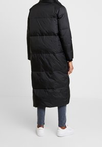 Svart, lång pufferjacka med horisontellt kviltade sektioner, hög krage och sidofickor. Bärs över jeans och vita sneakers.