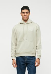 Γκρίζα φούτερ με κουκούλα και κορδόνια, μπροστινές τσέπες και ριμπ μανσέτες, με μικρό λευκό λογότυπο στο στήθος. Συνδυασμένο με μπλε τζιν.