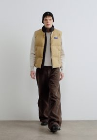 Beež värvi puffer-vest musta sisemise kihiga, kombineeritud kreemika soonikkoega kampsuni ja pruunide laiade pükstega. Must pipomüts ja tumedad jalatsid.