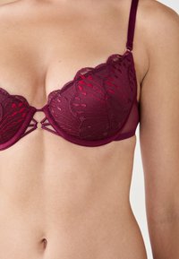 Bra in pizzo bordeaux con bordi scallopati, ricami floreali e dettaglio a apertura a chiave sul davanti. Spalline regolabili e ferretto morbido.