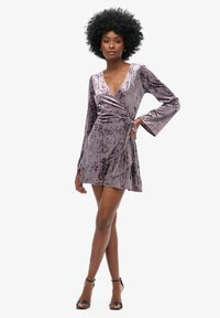 Fioletowa aksamitna sukienka otulająca z długimi rękawami, dekoltem w kształcie litery V, falbaną dolną częścią i dopasowaną sylwetką, zestawiona z czarnymi sandałami na cienkich obcasach.