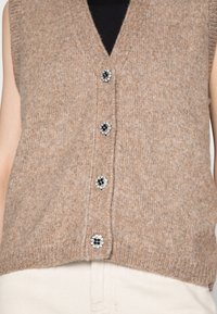 Gilet sans manches en tricot marron avec un profond col en V et cinq boutons décoratifs noirs ornés d'un motif floral.