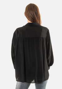 Blusa negra de manga larga con una textura suave y sedosa, que presenta un cuello con solapa y un detalle de pliegues en la parte posterior. Con un ajuste relajado.