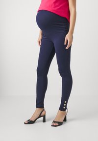 Marine gravide leggings med en glat tekstur, høj talje og tre knapdetaljer ved anklen, parret med sorte hæle-sandaler.