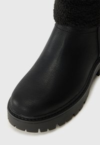 Botín de cuero negro hasta el tobillo con superficie texturizada, cuello acolchado y suela de goma gruesa con un prominente patrón de relieve.