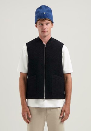 Zwarte wolvest met een ritssluiting aan de voorkant, twee zakken en een ronde halslijn, gelaagd over een wit t-shirt, met een blauwe gebreide muts.