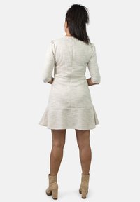 Vestido beige texturizado con falda acampanada, mangas tres cuartos y un cuerpo entallado, combinado con botines tobilleros de un tono marrón claro.