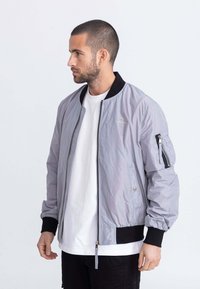 Giacca bomber grigio chiaro con polsini e colletto a coste neri, dotata di una zip frontale e di una tasca con cerniera sul maniche sinistro.