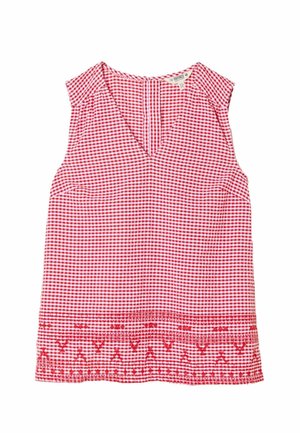 Frau mit langen braunen Haaren, die ein rot-weiß kariertes ärmelloses Top und marineblaue Shorts trägt, posiert mit den Händen in den Taschen vor einer schlichten Wand.