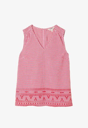 Frau mit langen braunen Haaren, die ein rot-weiß kariertes ärmelloses Top und marineblaue Shorts trägt, posiert mit den Händen in den Taschen vor einer schlichten Wand.