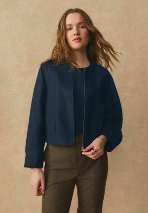 Világosbarna hajú nő sötét felső és olívazöld nadrág fölött tengerészkék cipzáras dzsekit visel, bézs háttér előtt, kezében a cipzár lehúzóját tartva.