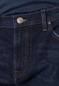 Mørkeblå denimjeans med en Lee-logo knapp, kontrasterende gul søm, og en klassisk passform. Har en jevn tekstur og sidelomme.