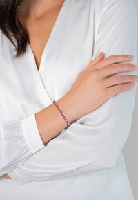 Sidabrinė apyrankė su pakaitomis violetiniais ir auksiniais karoliukais, dėvima ant riešo. Fone matoma balta, lygi medžiaga.