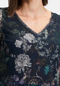 Vêtement à motif floral avec une base bleu marine, un décolleté en V et des nuances de vert, blanc et rose variées dans des designs botaniques complexes.