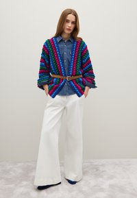Cárdigan de punto con textura de pompones multicolores en verde, azul y rosa, superpuesto a una camisa de mezclilla y combinado con pantalones anchos blancos.