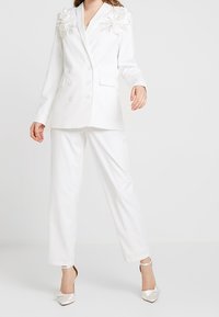 Biały, dopasowany żakiet z floralnym haftem na ramionach, podwójnie zapinany z przodu, zestawiony z dopasowanymi spodniami i szpilkami w szpic.