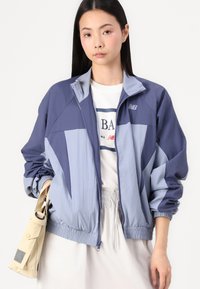 Navy and light blue nylon windbreaker jacket with a zip front, elastic cuffs, and hem. Features color-blocked design and New Balance logo.