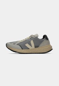 Sporta sneakers ar pelēku tīkla augšdaļu, bēšas gumijas vidusdaļu un melnu papēdi. Iezīmē krēmkrāsas šņores un lielu bēšu logo sānos.