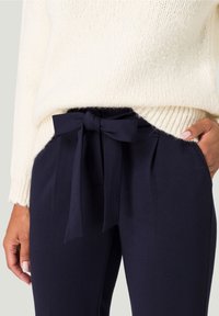 Gros plan sur une personne portant un pantalon bleu marine avec une ceinture en tissu nouée en nœud et un pull en tricot crème, la main reposant dans la poche.