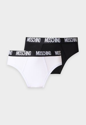 Homme torse nu, debout pieds nus en slip blanc avec une ceinture noire estampillée "Moschino" sur un fond clair uni.