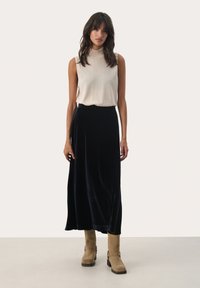 Top senza maniche beige abbinato a una lunga gonna in velluto blu navy e stivaletti color tan con dettagli in fibbia, in piedi su uno sfondo neutro.