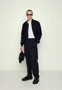 Μαύρο bomber μπουφάν με φερμουάρ, λευκό t-shirt, σκούρο παντελόνι, μαύρα sneakers και μια μικρή μαύρη τσάντα. Οι καθαρές γυαλιά ηλίου προσθέτουν έμφαση στην εμφάνιση.